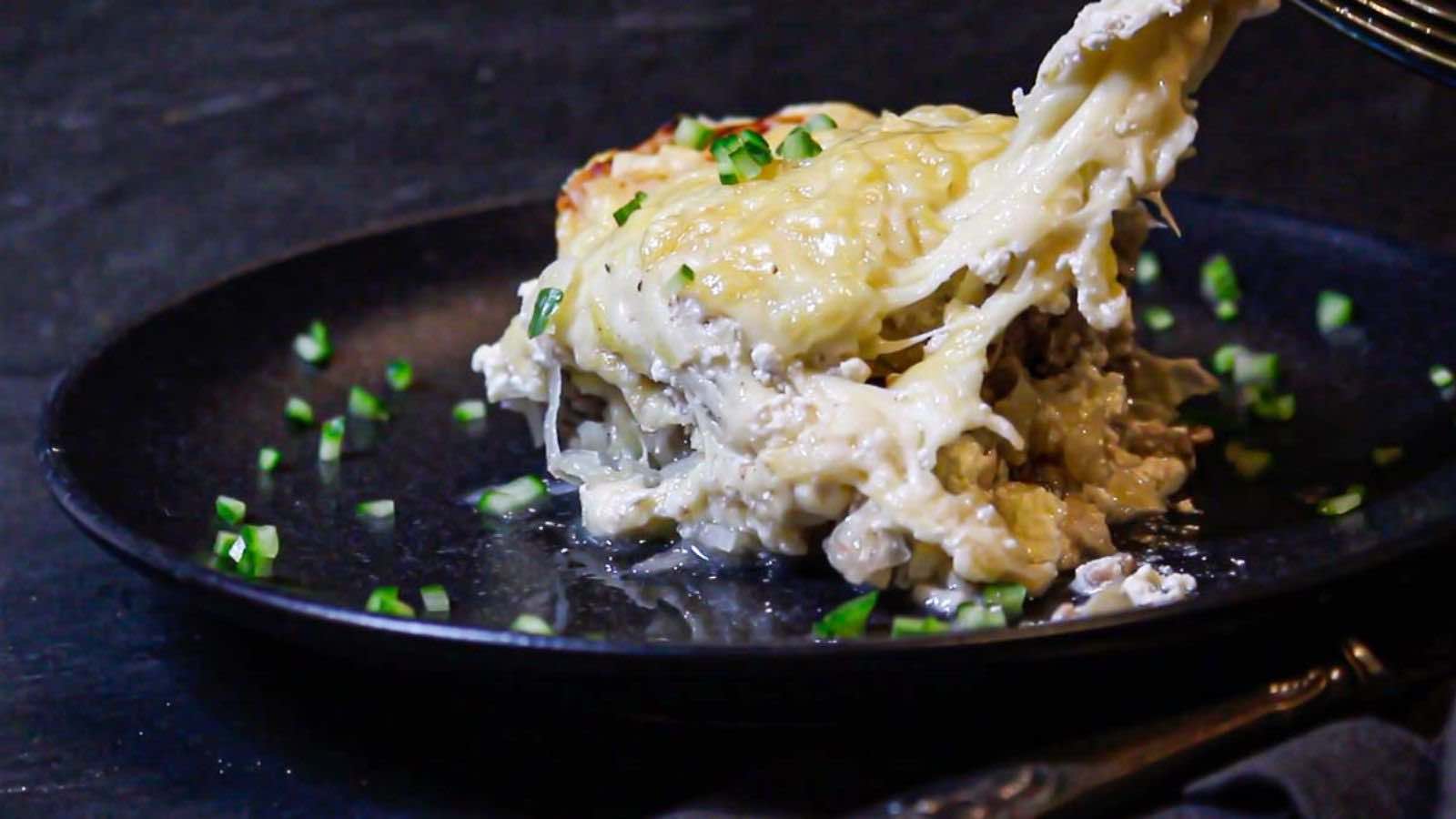 A serving of cheesy lasagna being lifted from a dark plate, garnished with chopped green herbs.