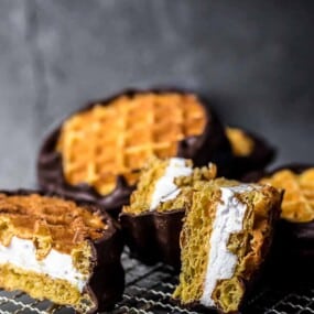 Waffle sandwiches filled with cream and partially dipped in chocolate are arranged on a wire cooling rack.