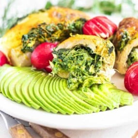 A white plate with sliced avocado, stuffed chicken with spinach, and three whole radishes.