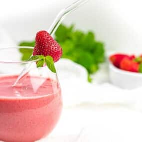 A glass of strawberry smoothie with a glass straw, garnished with a whole strawberry. A bowl of strawberries and some green foliage are in the background.