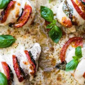Four baked chicken breasts stuffed with cheese, tomatoes, and basil, garnished with fresh basil leaves on a baking tray.