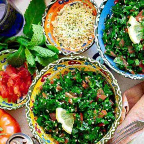 A table setting with bowls of tabbouleh, chopped tomatoes, herbs, utensils, and a glass of water. A lemon and tomato lie nearby.