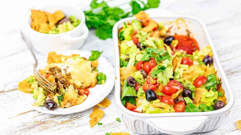A serving of taco salad in a white rectangular dish and a small portion on a white plate with a fork. The salad contains lettuce, tomatoes, black olives, and tortilla chips on a white wooden surface.
