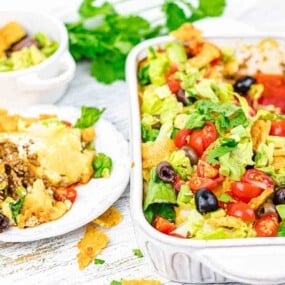 A serving of taco salad in a white rectangular dish and a small portion on a white plate with a fork. The salad contains lettuce, tomatoes, black olives, and tortilla chips on a white wooden surface.