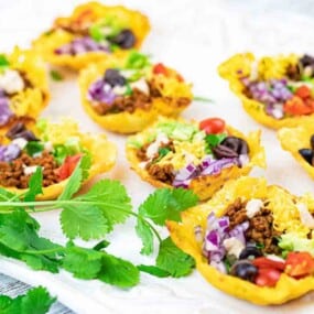 Mini taco bites filled with ground meat, tomatoes, shredded lettuce, cheese, olives, and purple cabbage, served on a white platter with fresh cilantro garnish.
