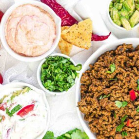 A spread of taco ingredients includes a bowl of cooked ground beef, chopped herbs, sliced avocado, a bowl of pink sauce, tortilla chips, grape tomatoes, and a small bowl of creamy dip.