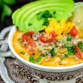 A bowl of soup topped with avocado slices, shredded cheese, cherry tomatoes, cilantro, and a lime wedge, served on a patterned plate.