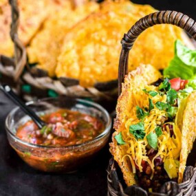 A basket of tacos filled with lettuce, cheese, and meat sits next to a bowl of salsa on a dark cloth. More tacos are visible in a basket in the background.