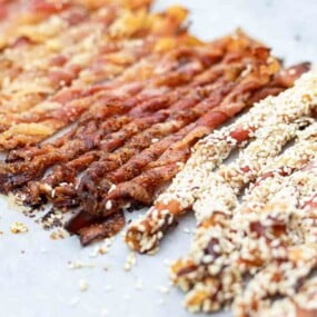 A close-up of cooked bacon strips, some plain and some coated with sesame seeds, laid out on a white surface.