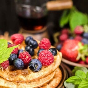 A close-up of waffles topped with blueberries, raspberries, and a green leaf. A plate of mixed berries and a container of syrup are in the background.