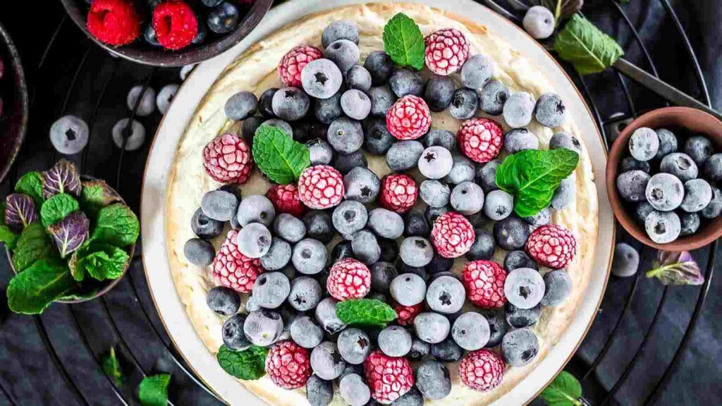 A cheesecake topped with a mix of frozen blueberries and raspberries, garnished with mint leaves. Bowls of extra berries and mint leaves are nearby.
