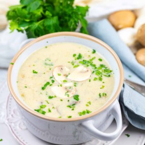 A bowl of creamy mushroom soup garnished with chopped parsley and sliced mushrooms, placed on a white plate next to a blue napkin and a bunch of fresh parsley.