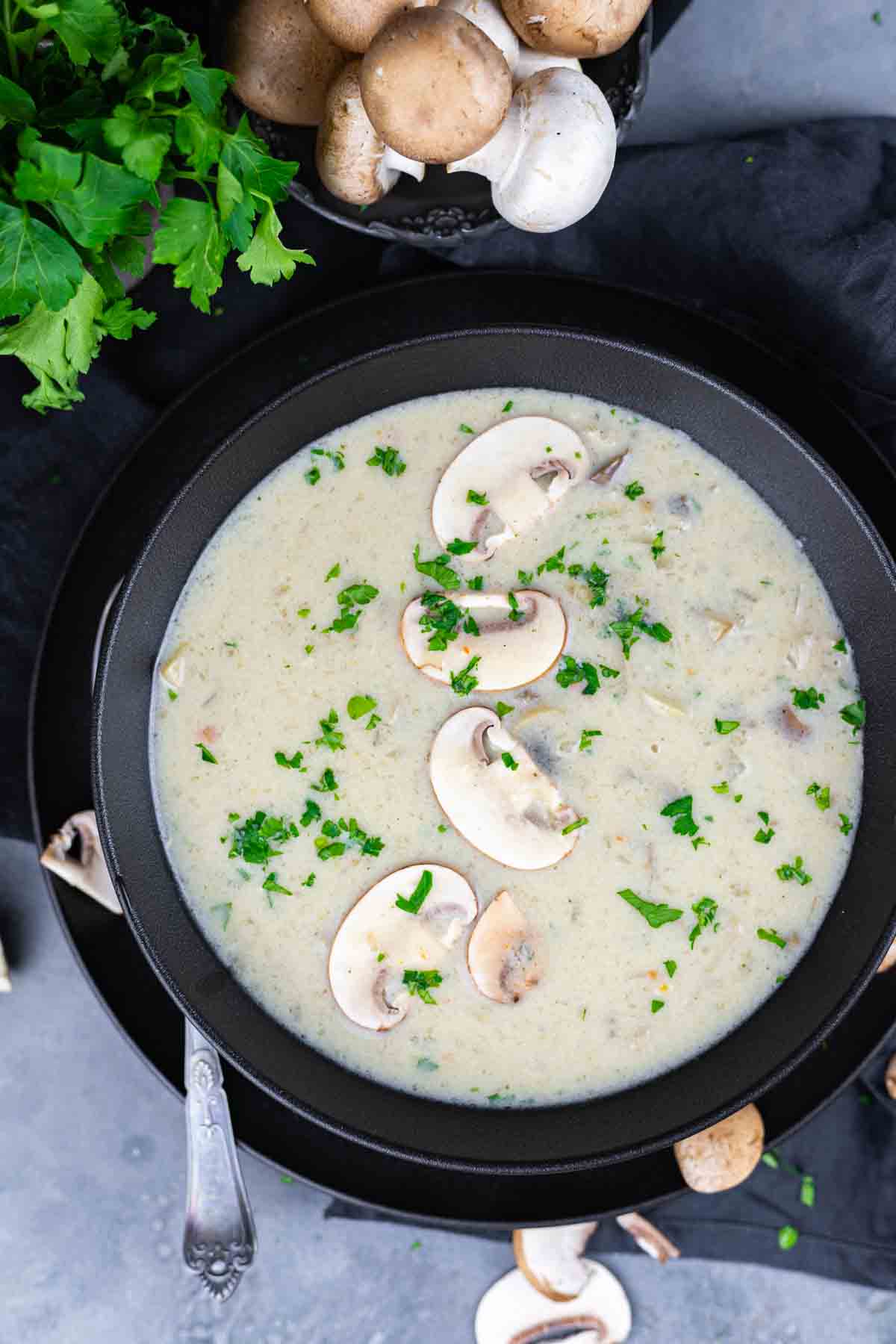 A black bowl filled with creamy mushroom soup, garnished with sliced mushrooms and chopped parsley. Fresh mushrooms and parsley are placed around the bowl on a dark surface.