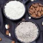 Two bowls of ground almonds, one finer and one coarser, with whole almonds and almond pieces scattered around on a dark fabric background.