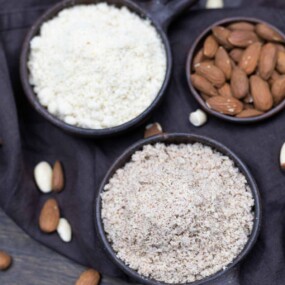 Two bowls of ground almonds, one finer and one coarser, with whole almonds and almond pieces scattered around on a dark fabric background.