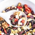 A white spoon filled with a variety of trail mix ingredients, including nuts, seeds, dried fruits, and chocolate chips, held over a bowl containing the same mixture.