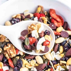 A white spoon filled with a variety of trail mix ingredients, including nuts, seeds, dried fruits, and chocolate chips, held over a bowl containing the same mixture.