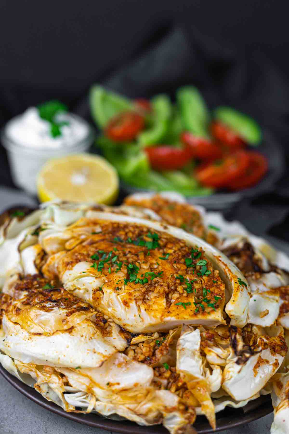 A plate of baked cabbage topped with seasonings, garnished with green herbs. In the background, there is a side salad with tomatoes and a small bowl of sour cream.