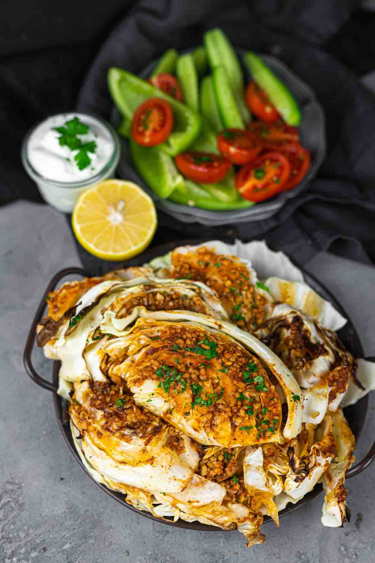 A plate of roasted cabbage garnished with herbs, accompanied by a side of cucumber and tomato slices, half a lemon, and a small bowl of white sauce topped with green garnish.