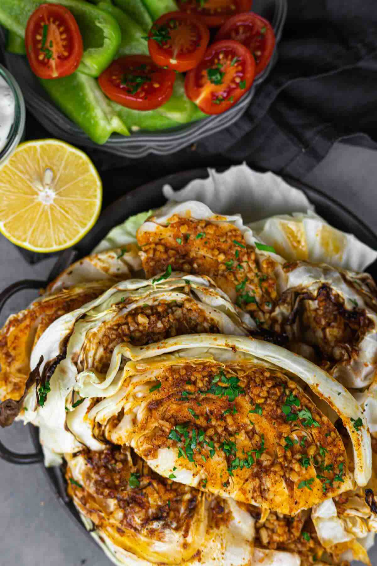 Grilled cabbage wedges seasoned with spices and herbs, garnished with chopped parsley, served alongside tomato and pepper salad and a halved lemon.