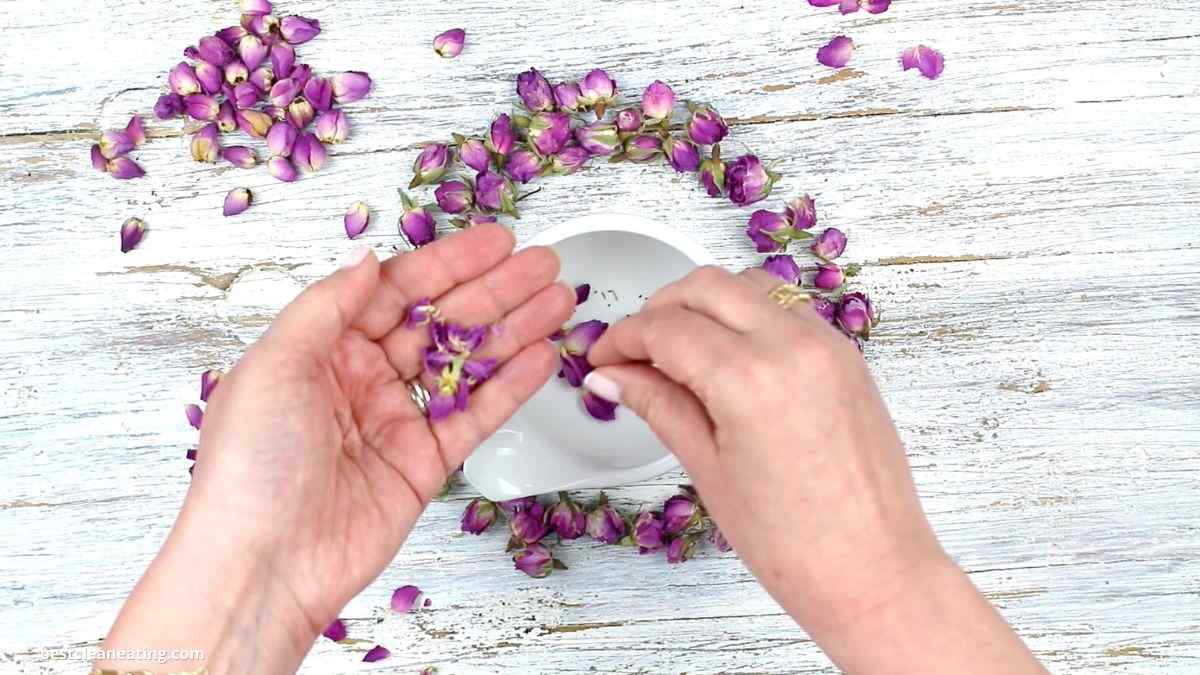 Hands arranging a wreath of small purple flowers on a white wooden surface, with a white plate at the center. Additional flowers are scattered around.