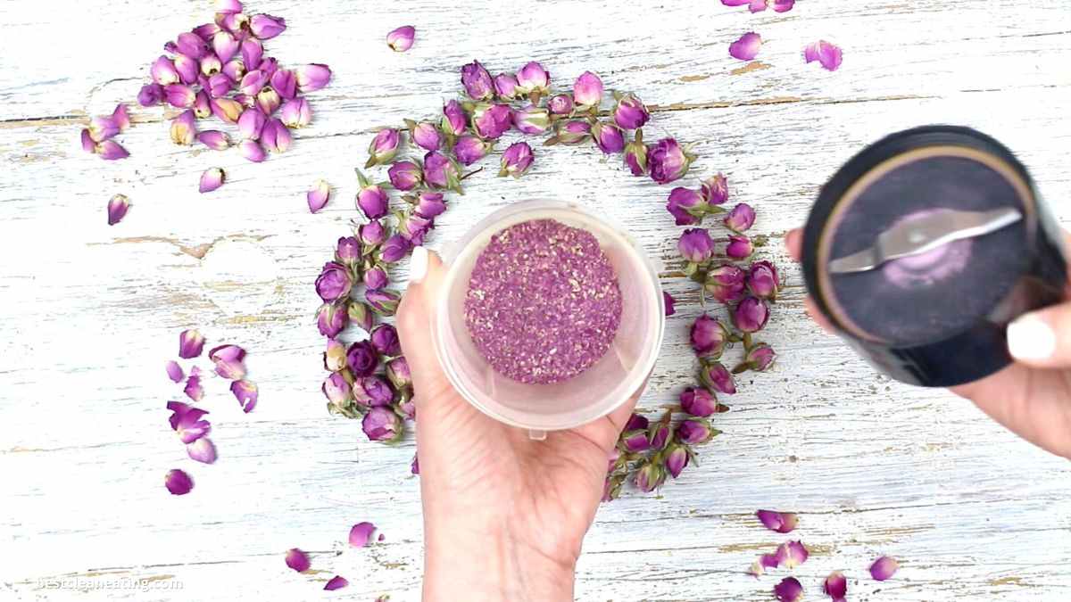 Close-up of hands holding a blender with crushed purple flower petals in it. Dried flower buds and petals are scattered on a white wooden surface, forming a partial circle around the blender.