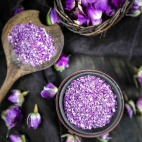 A small bowl and a wooden spoon filled with purple rose powder sit next to a wicker basket containing purple rose petals, all arranged on a dark fabric surface.