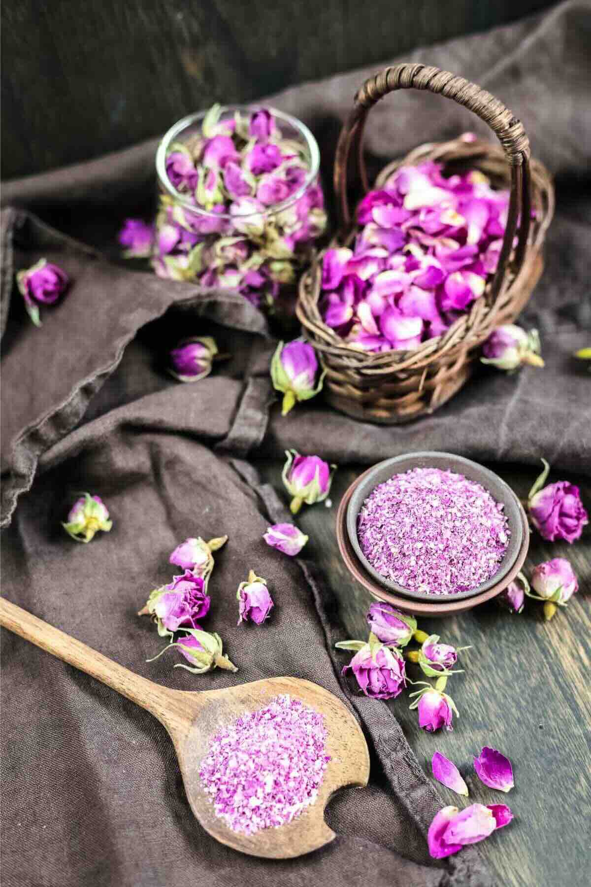 A wooden table holds a basket of pink rose petals, a bowl of crushed petals, a spoon with petal fragments, and some loose rosebuds, all set on a dark cloth.