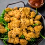 A dish of fried chicken bites served on a bed of arugula with a small bowl of dipping sauce in the background.