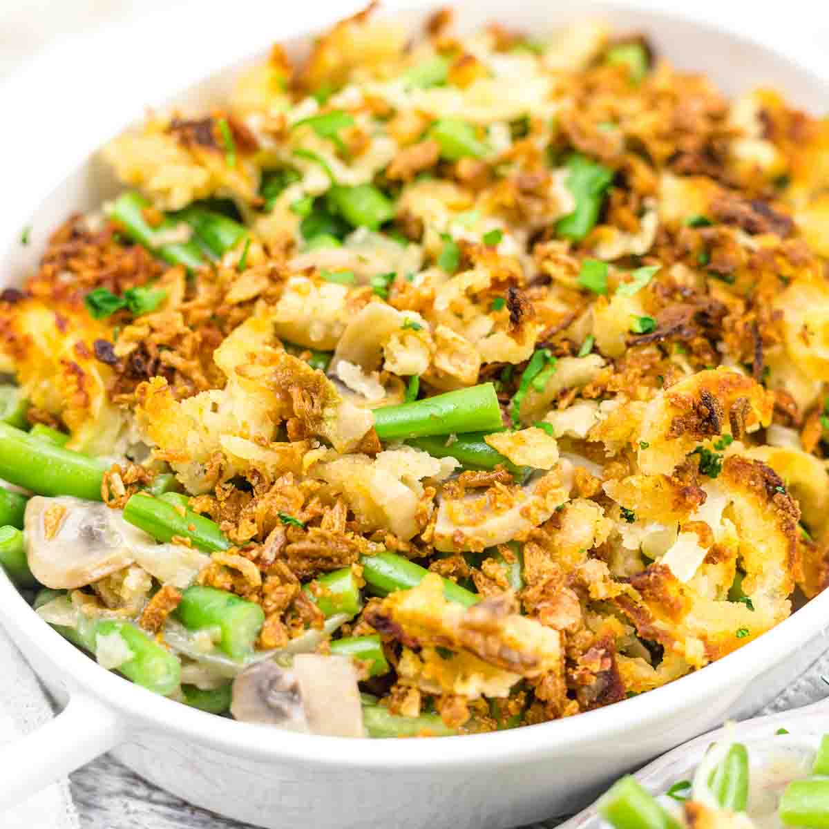A close-up of a baked casserole in a white dish, containing green beans, mushrooms, crispy fried onions, and a breadcrumb topping.