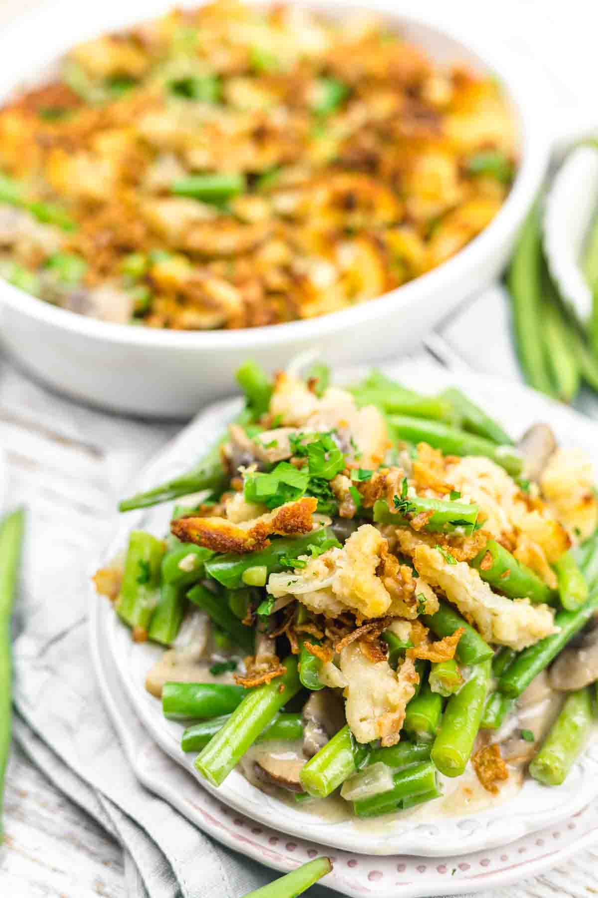 A plate of green bean casserole topped with crispy onions. Another dish of the same casserole is in the background. Green beans and a napkin are on the side.