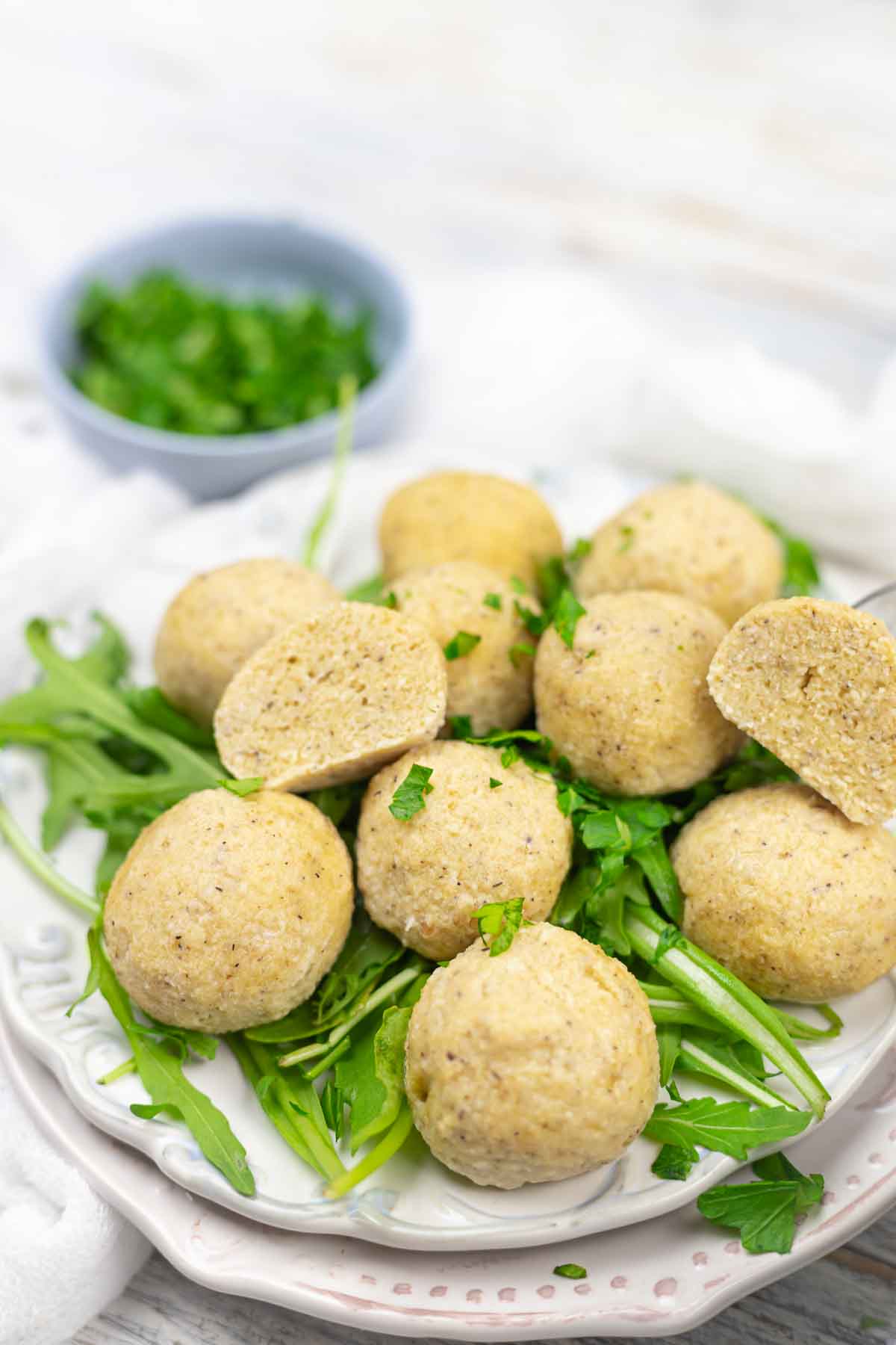 Plate of round, cooked dumplings on a bed of fresh greens, garnished with chopped herbs. A halved dumpling is displayed at the top. A bowl of herbs is in the background.