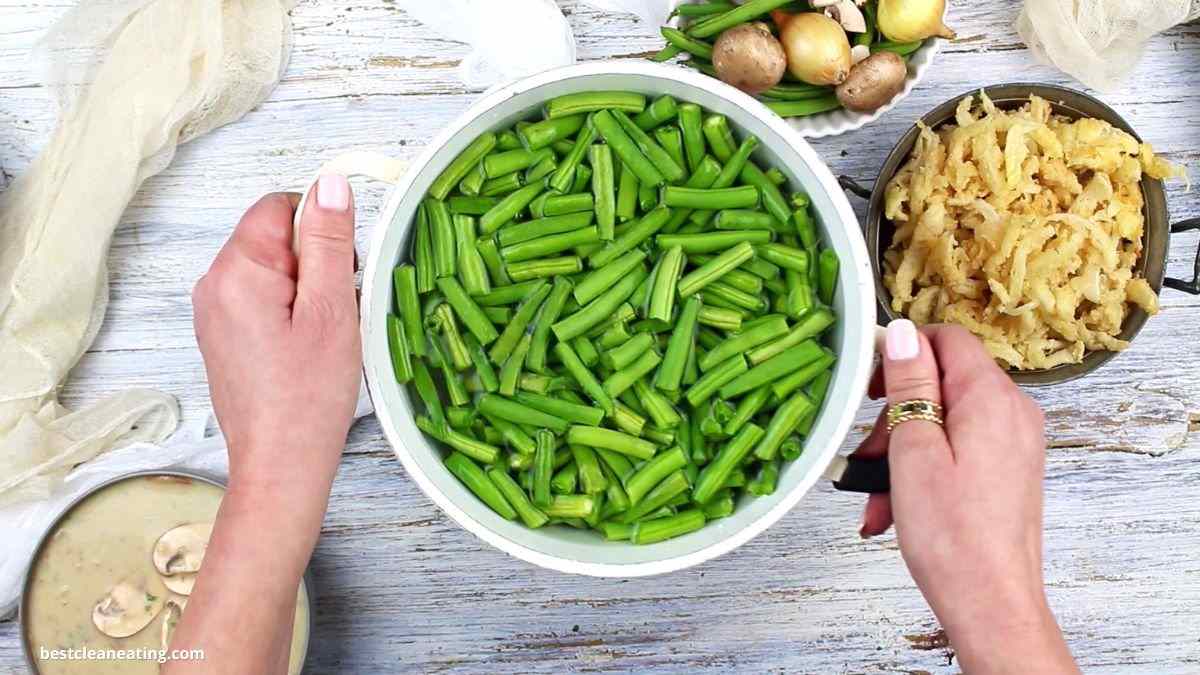 Hands holding a pot of sliced green beans, surrounded by mushrooms and shredded vegetables on a light wooden surface.