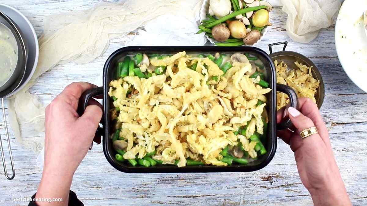 Hands holding a baking dish with a casserole topped with crispy onions, green beans, and mushrooms. Nearby are a bowl of small potatoes and a saucepan.