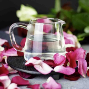 A glass pitcher of clear liquid on a black plate, surrounded by pink and white rose petals.