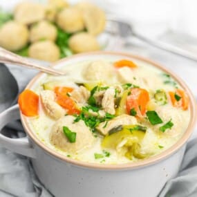 A white bowl filled with creamy chicken soup, featuring chunks of chicken, meatballs, sliced carrots, zucchini, and sprinkled with fresh parsley.