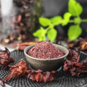 A small bowl filled with ground hibiscus sits on a black surface, surrounded by dried hibiscus petals and fresh green leaves in the background.