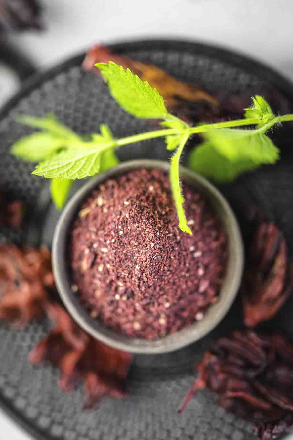 A small bowl of purple-hued spice on a dark textured surface, with a sprig of green leaves placed beside it. Dried petals are scattered around the bowl.