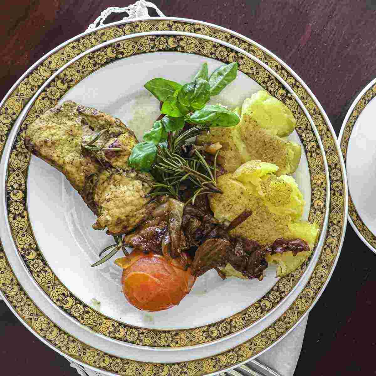 A plate with pork fillet, herb garnish, roasted potatoes, and a small roasted tomato, on a decorative plate.
