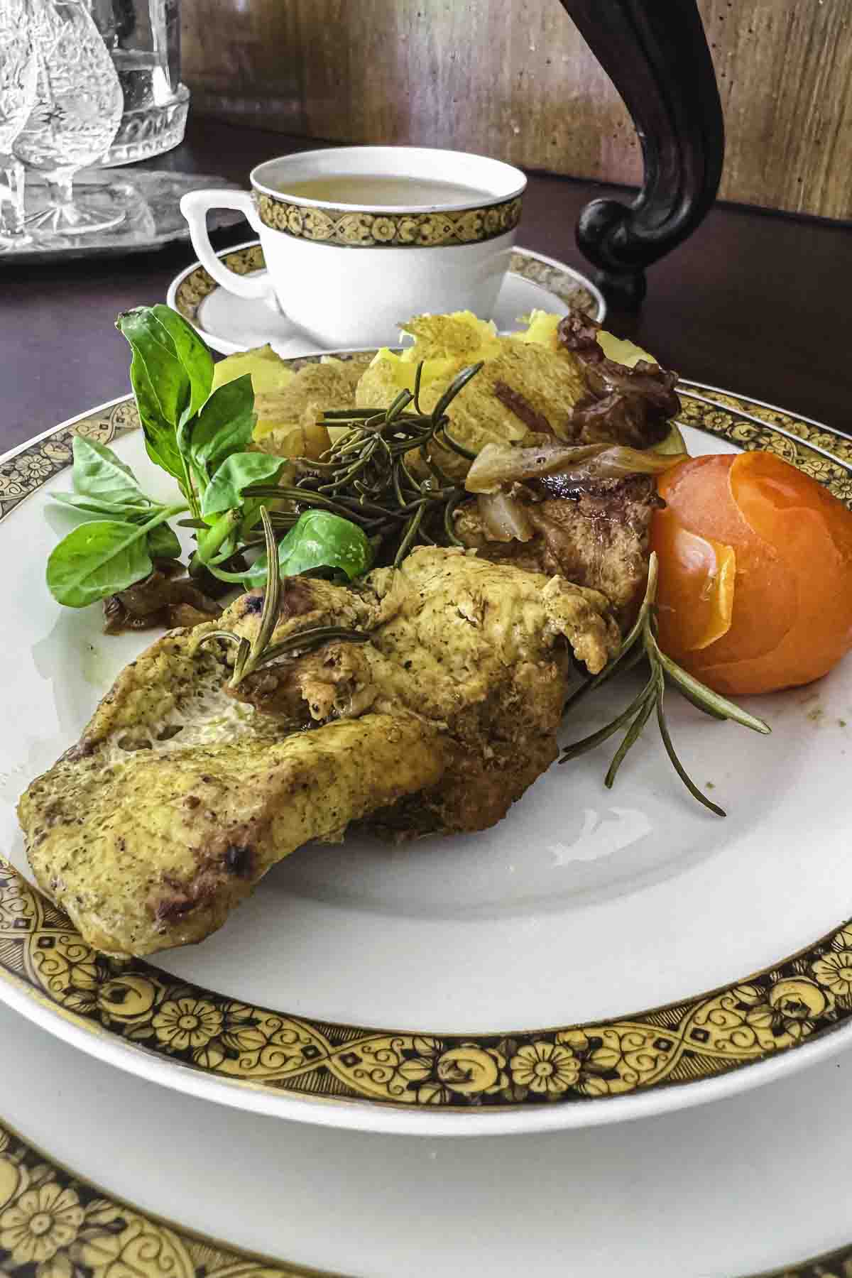 A plate of pork fillet with greens, sliced tomato, and garnish sits beside a decorative cup on an ornate dish.