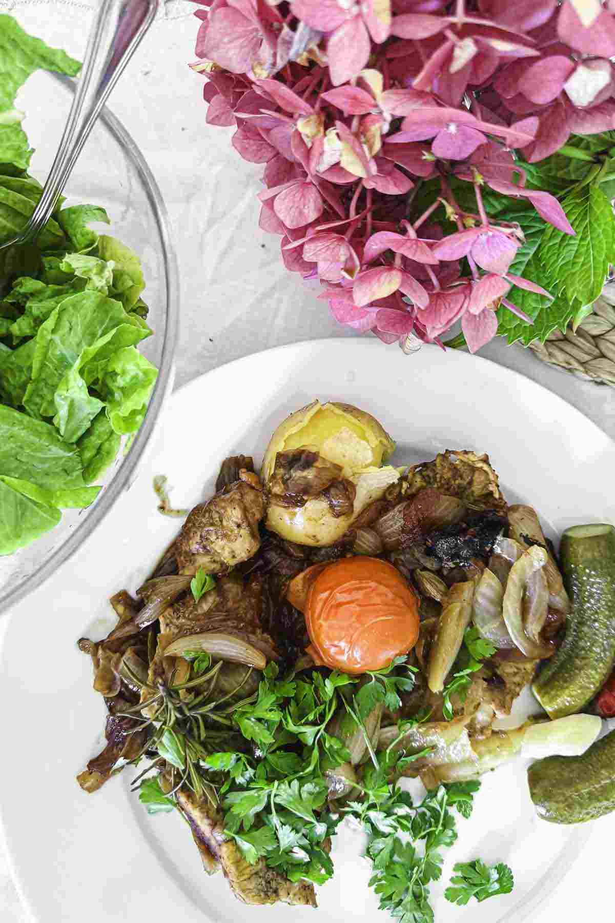 A plate of roasted vegetables and herbs with a small bowl of green salad beside it. Pink hydrangea flowers are visible in the background.