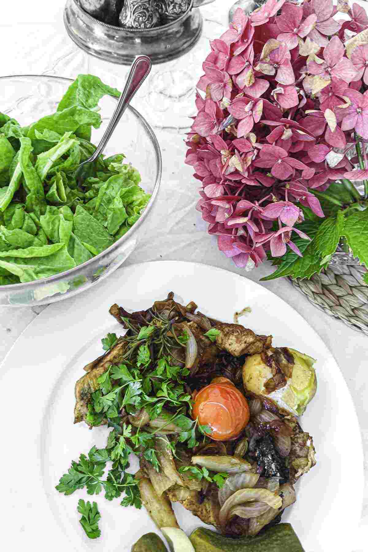 A plate of pork fillet with greens and a cherry tomato, next to a bowl of leafy salad and a bouquet of pink hydrangeas.