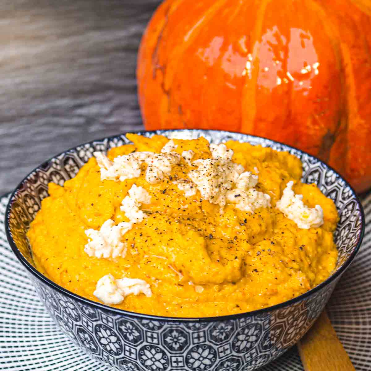A bowl of mashed pumpkin, reminiscent of delightful rice cauliflower recipes, is topped with crumbled white cheese and set on a patterned tablecloth, with a whole pumpkin in the background.