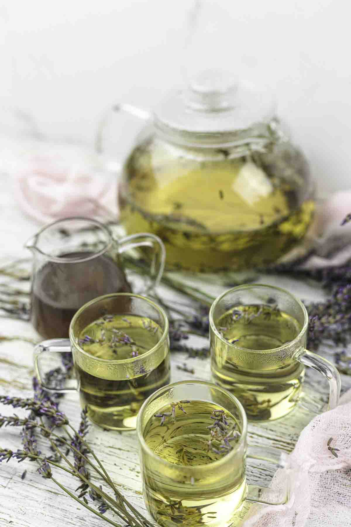 Glass teapot and three cups filled with greenish herbal tea, garnished with lavender sprigs, on a white wooden surface. Lavender buds scattered around.
