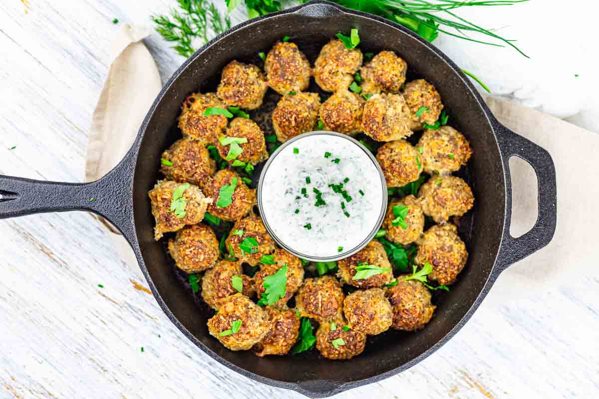 Cast iron skillet with cream cheese sausage balls arranged around a bowl of white dip, garnished with chopped herbs.