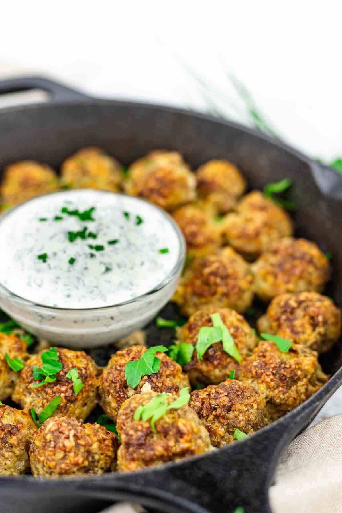 A skillet filled with browned meatballs, garnished with parsley, surrounding a small bowl of creamy sauce.
