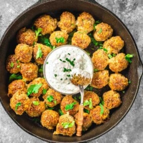 A skillet filled with browned meatballs, garnished with herbs, surrounding a small bowl of creamy dip with a spoon.