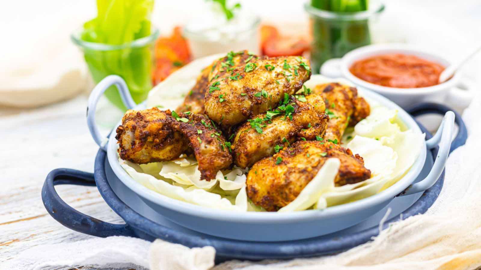 A plate of seasoned, chicken pieces garnished with herbs, served on cabbage leaves, with small bowls of sauce and vegetables in the background.
