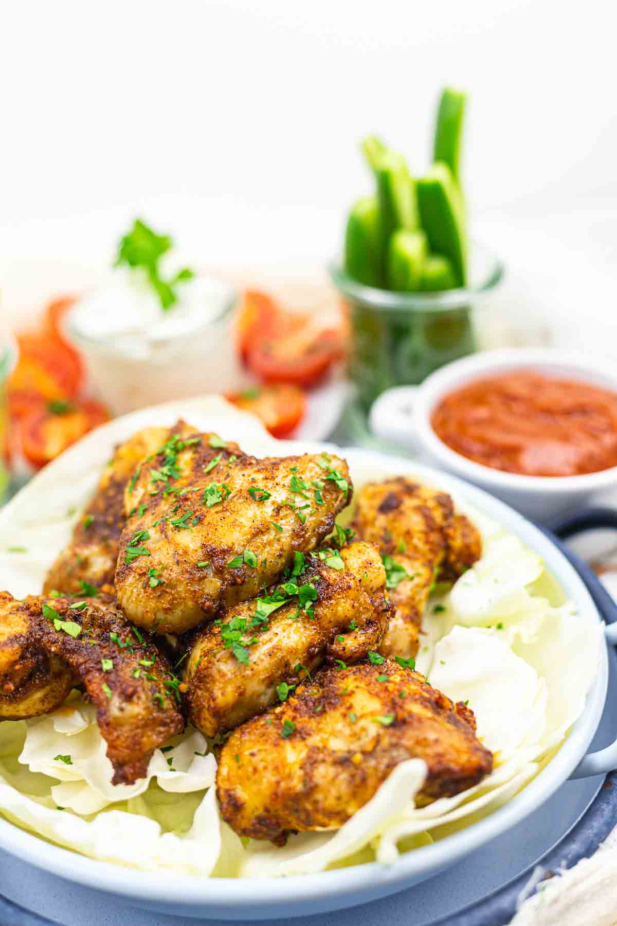 A bowl of seasoned chicken wings garnished with herbs, served on cabbage. Accompanied by dips and sliced vegetables in the background.