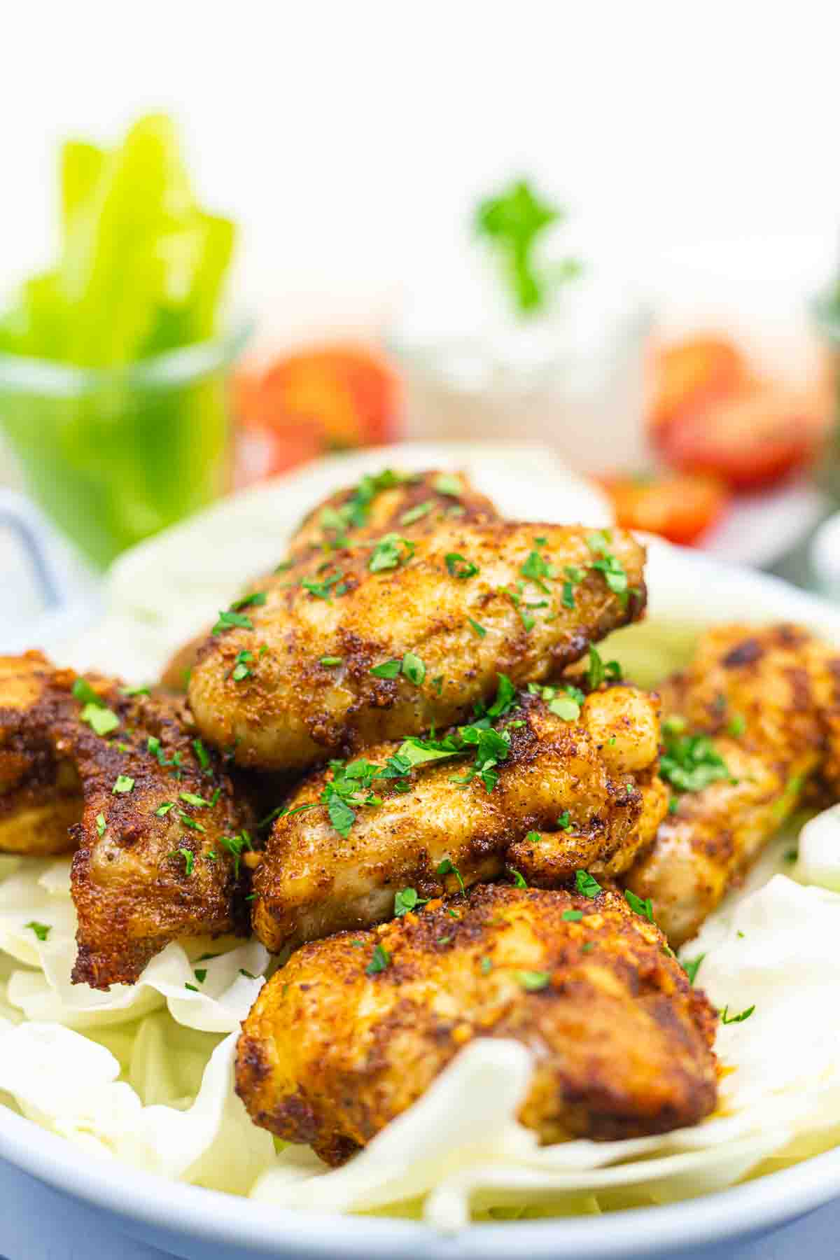 A plate of seasoned, grilled chicken wings garnished with chopped herbs, served on a bed of lettuce, with blurred vegetables in the background.
